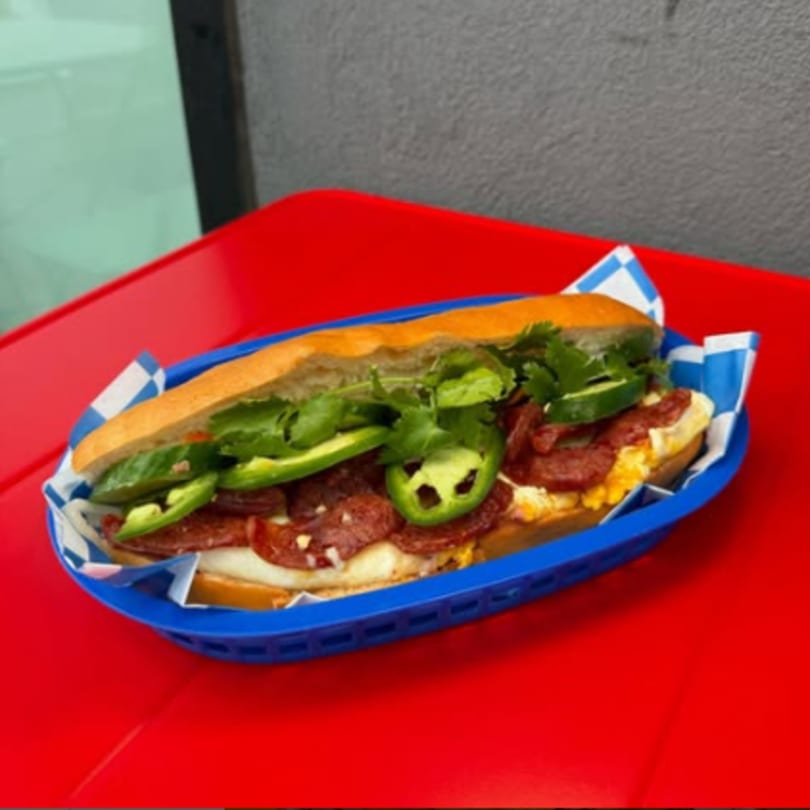 A Jersey Banh Mi sandwich with avocado, cilantro, and jalapeños in a blue basket on a red table.