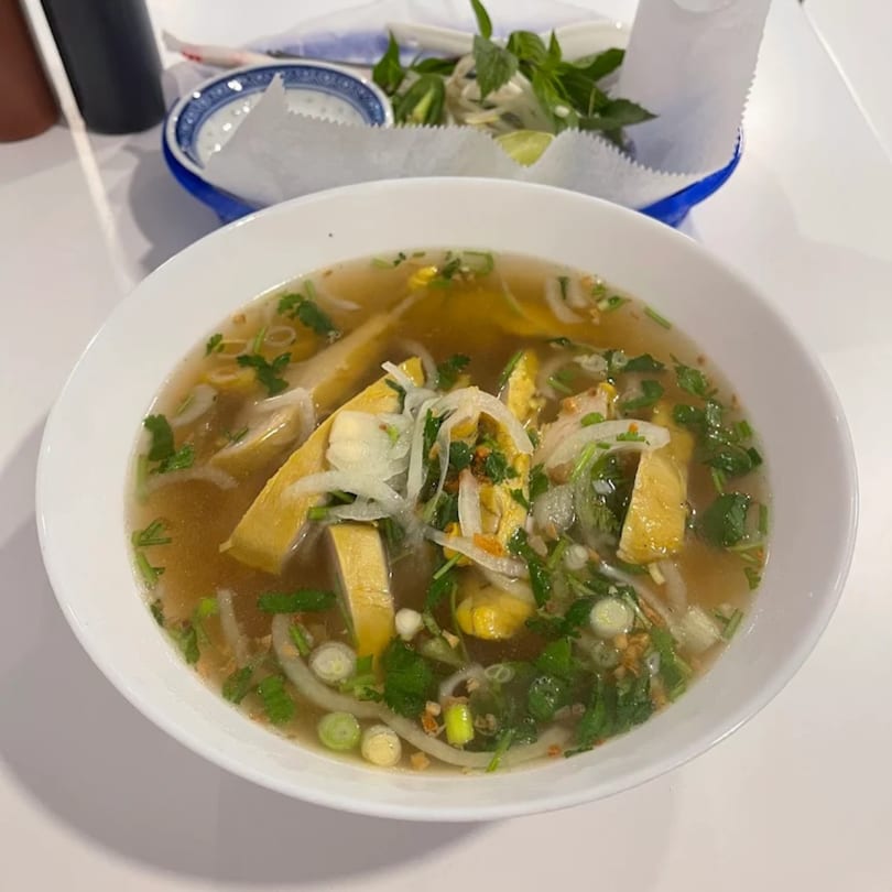 A bowl of Vietnamese pho with tofu, onions, and herbs, with a side of fresh basil.